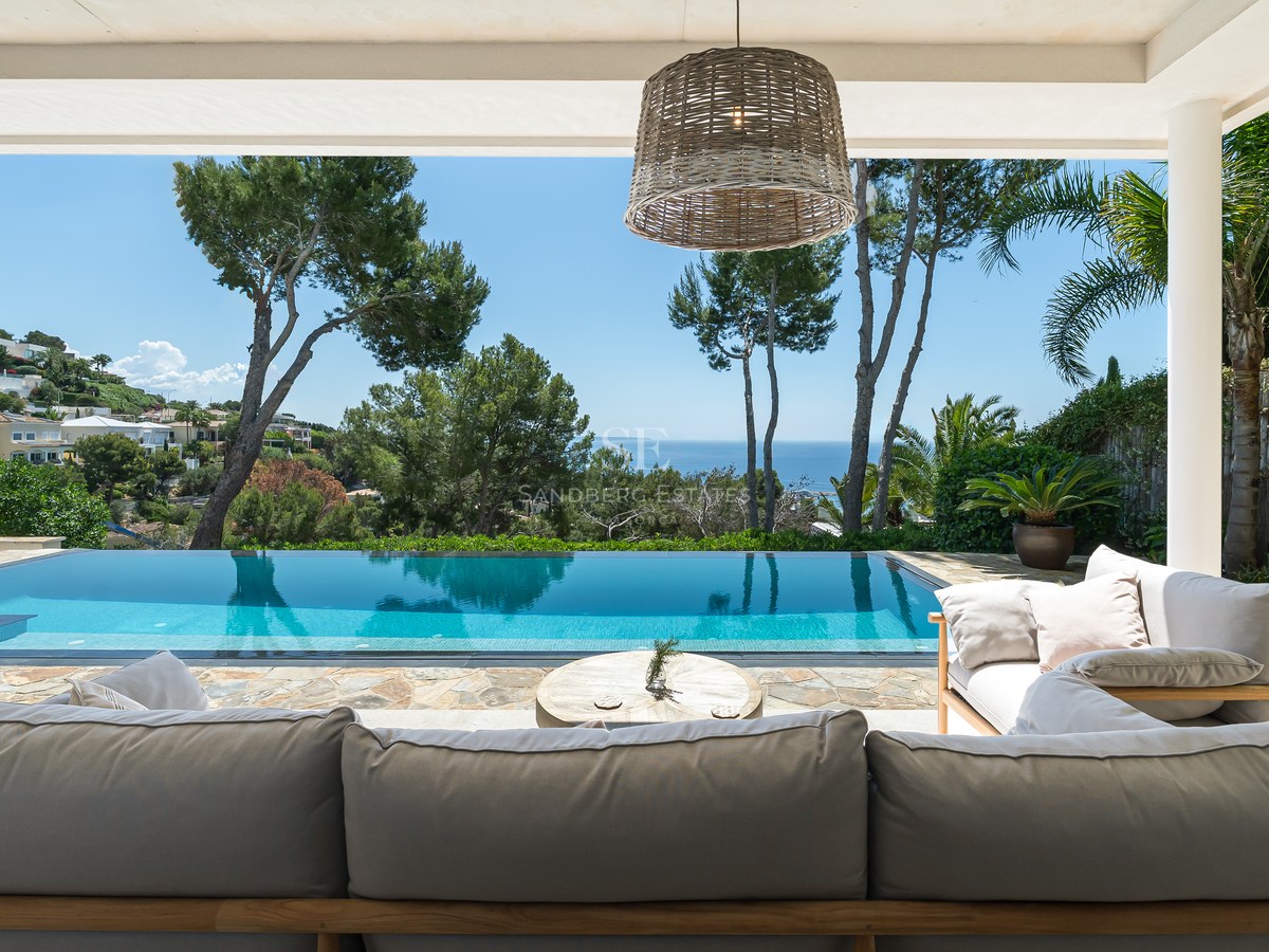 Infinitypool och loungegrupp under en skuggad terrass med utsikt över havet och lummiga tallar.