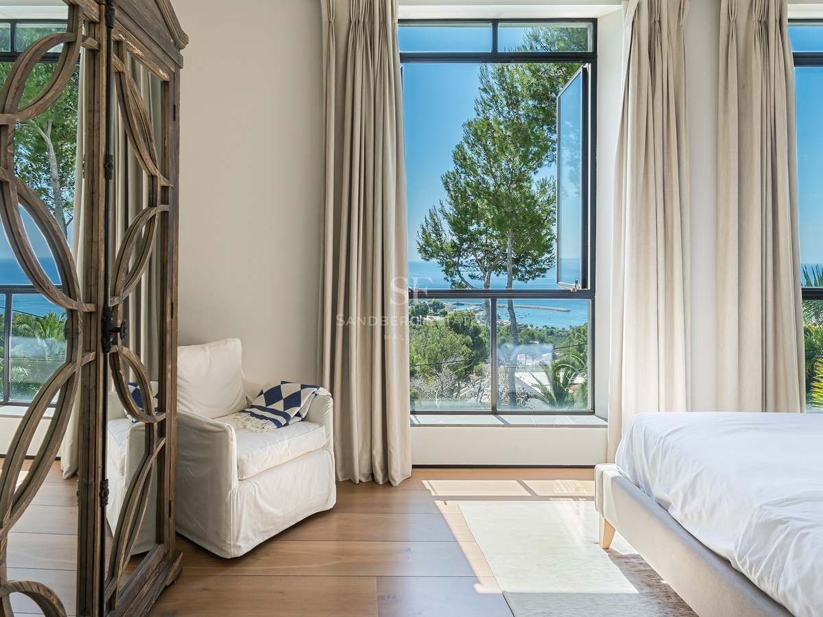 Ljust master bedroom med stora fönster mot havet, en vit fåtölj och en utsmyckad garderob i trä.