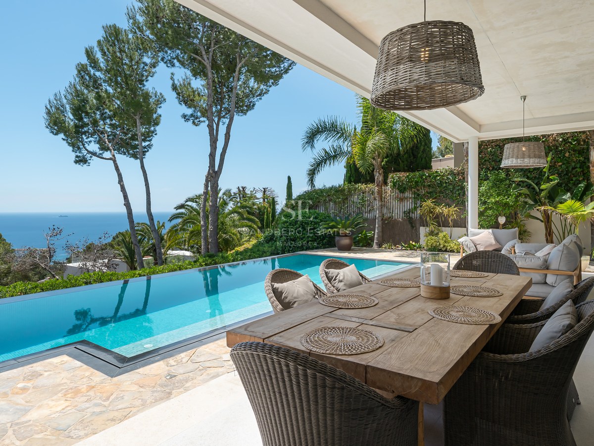Terrass med matbord i trä bredvid en infinitypool med utsikt över blått hav och tallar.