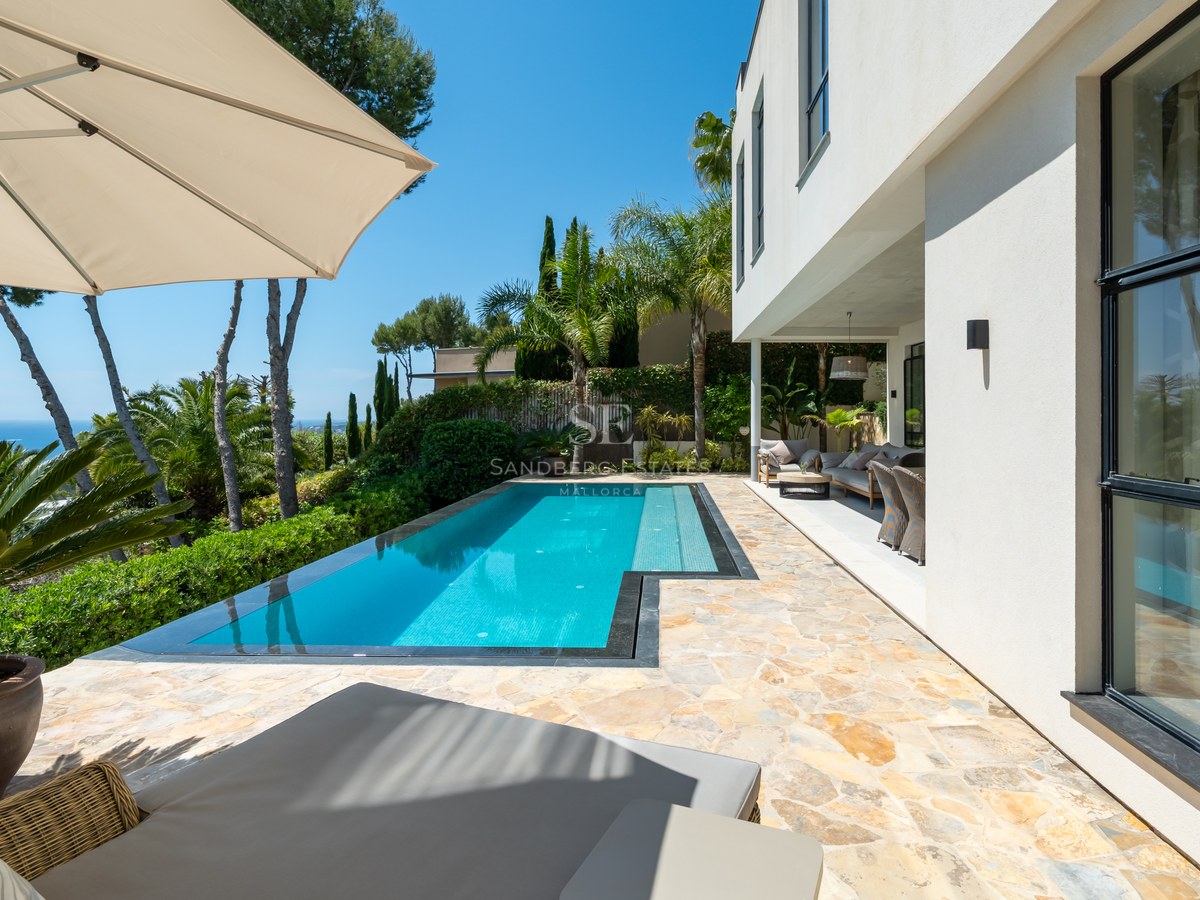 En turkos pool på en stenterrass bredvid en vit modern villa med utsikt över havet.