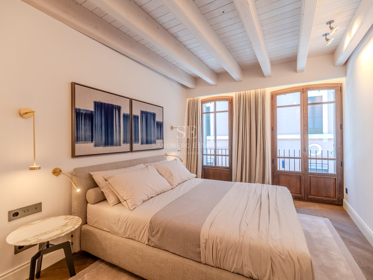 Chambre de luxe avec plafonds à poutres blanches, portes-fenêtres en bois et appliques en laiton.