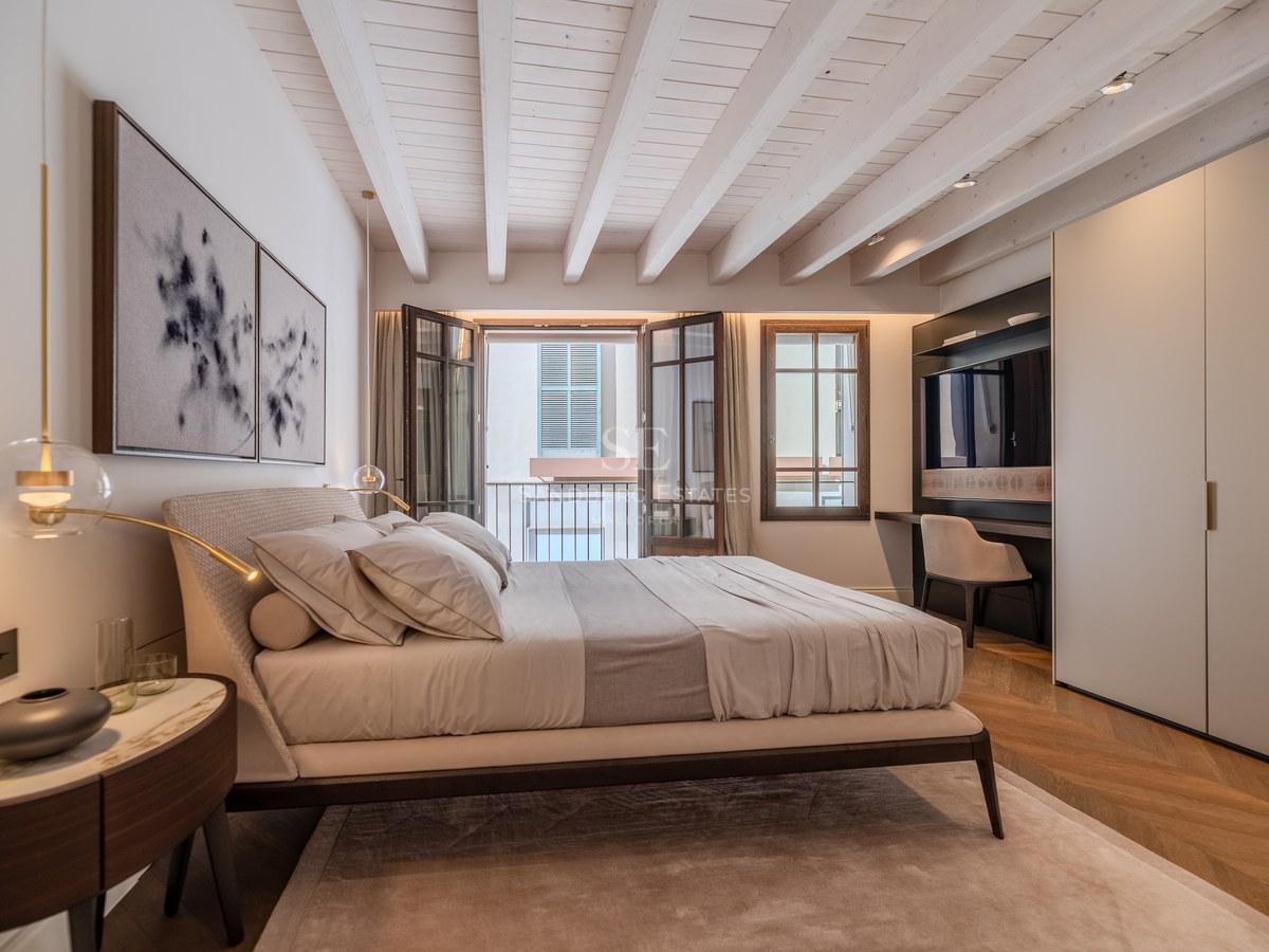 Chambre élégante avec plafond à poutres blanches, parquet en point de Hongrie et balcon donnant sur une rue.