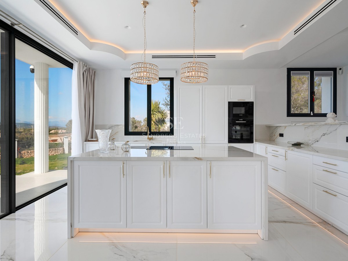 Cocina moderna y luminosa con gran isla de mármol, detalles dorados y ventanales de suelo a techo hacia el jardín.