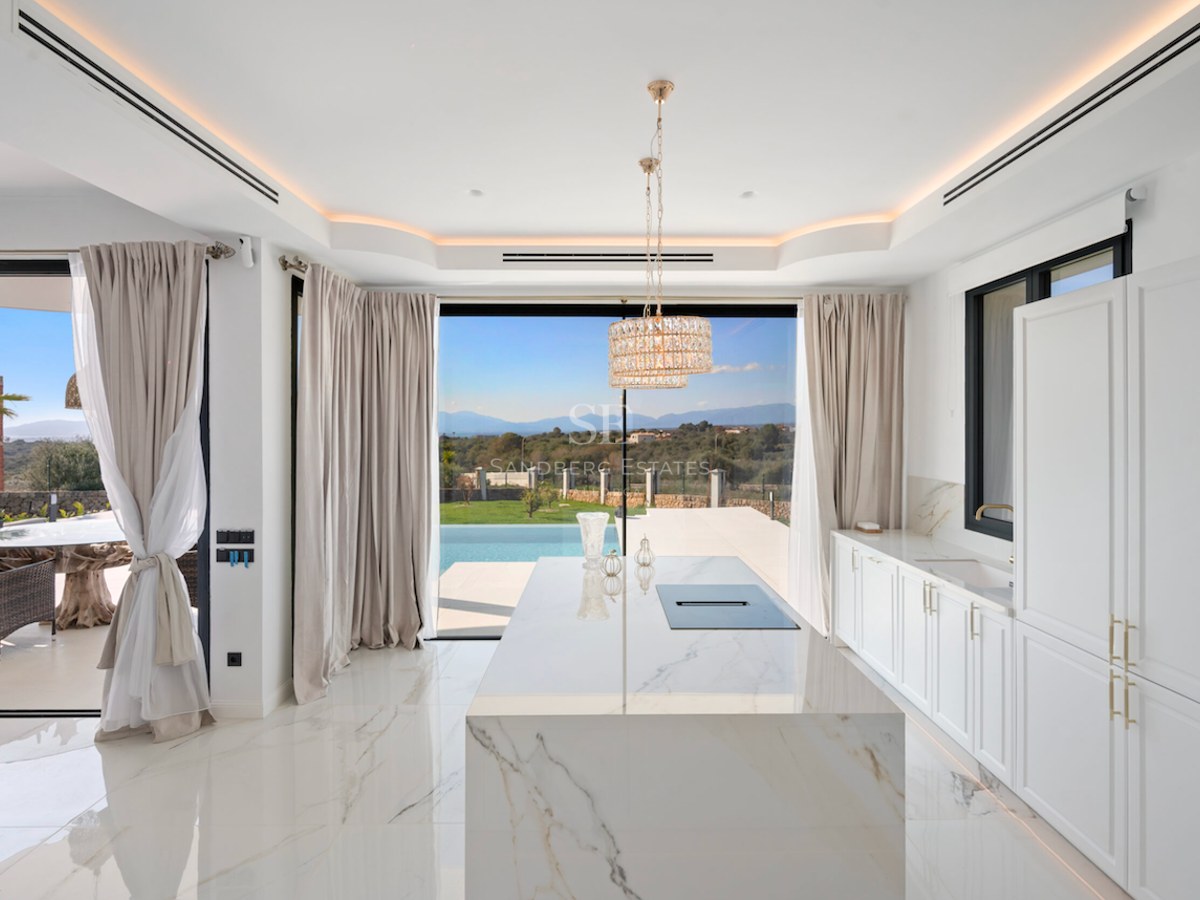 Cocina moderna con gran isla de mármol blanco, muebles blancos y grandes ventanales con vistas a la piscina y el paisaje.