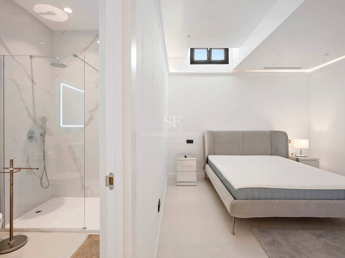Dormitorio moderno con cama gris y puerta abierta hacia una ducha de mármol con mampara de cristal.