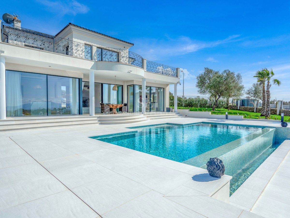 Villa blanca de estilo mediterráneo moderno con piscina infinita, terraza de piedra clara y jardín bajo cielo azul.