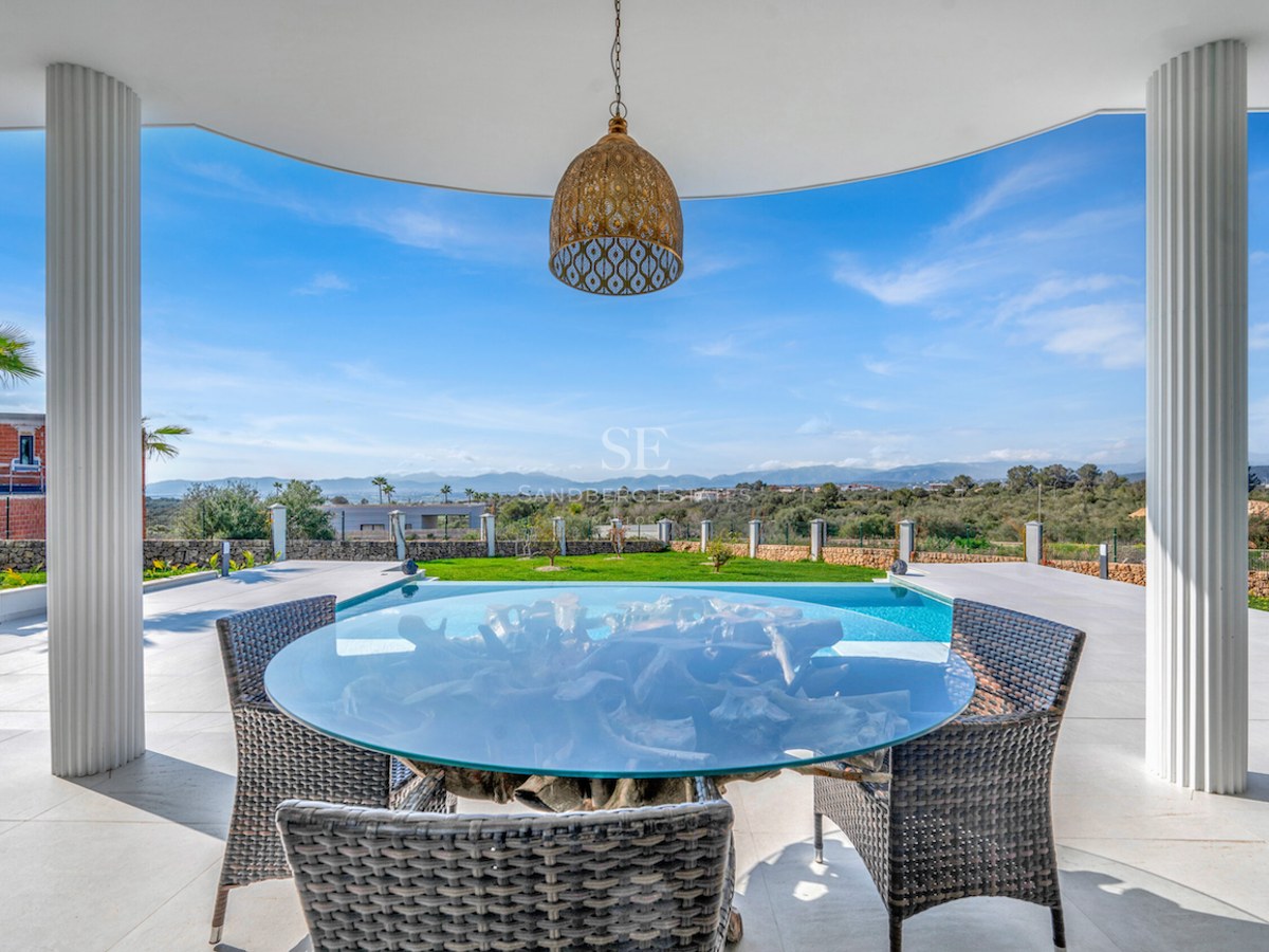 Terraza cubierta con mesa de cristal y sillas de mimbre frente a una piscina con vistas a la montaña.