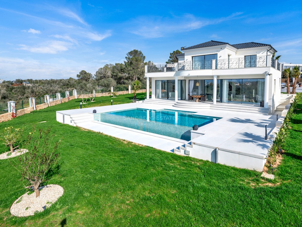 Vista de una villa blanca de dos plantas con piscina integrada, terraza de piedra y amplio jardín bajo cielo azul.