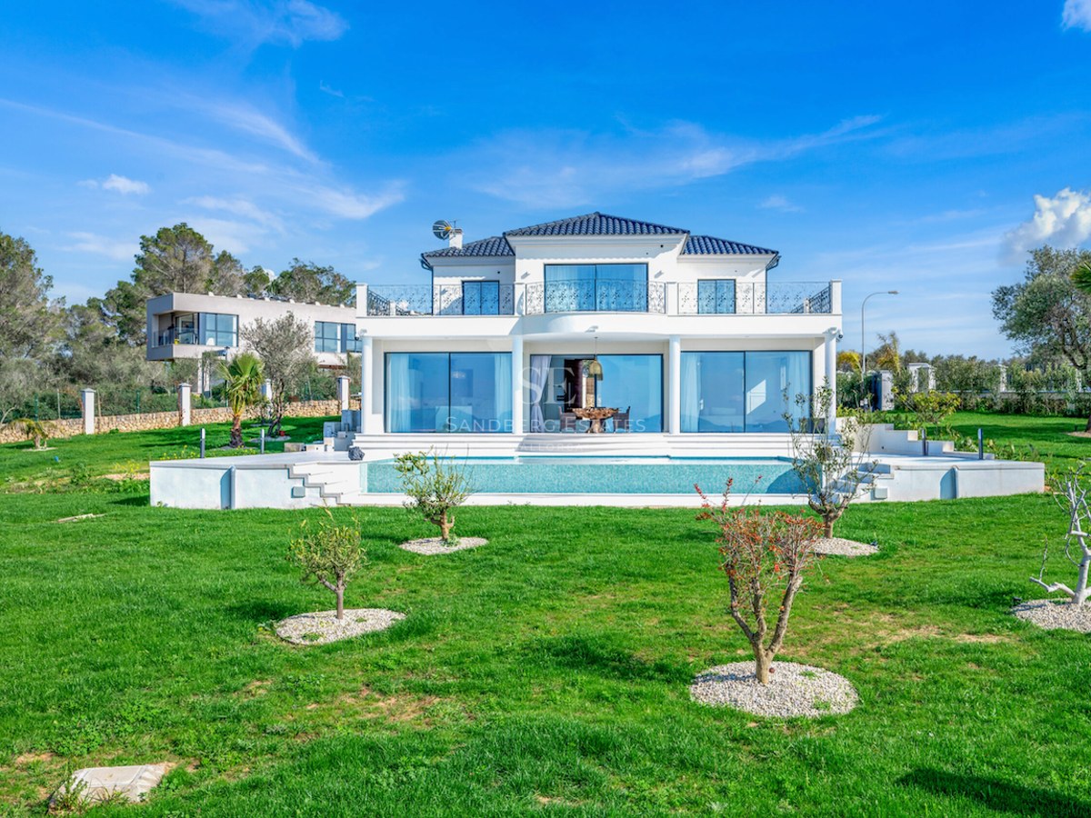 Fachada frontal de una villa de lujo blanca con piscina y amplio jardín bajo un cielo despejado.