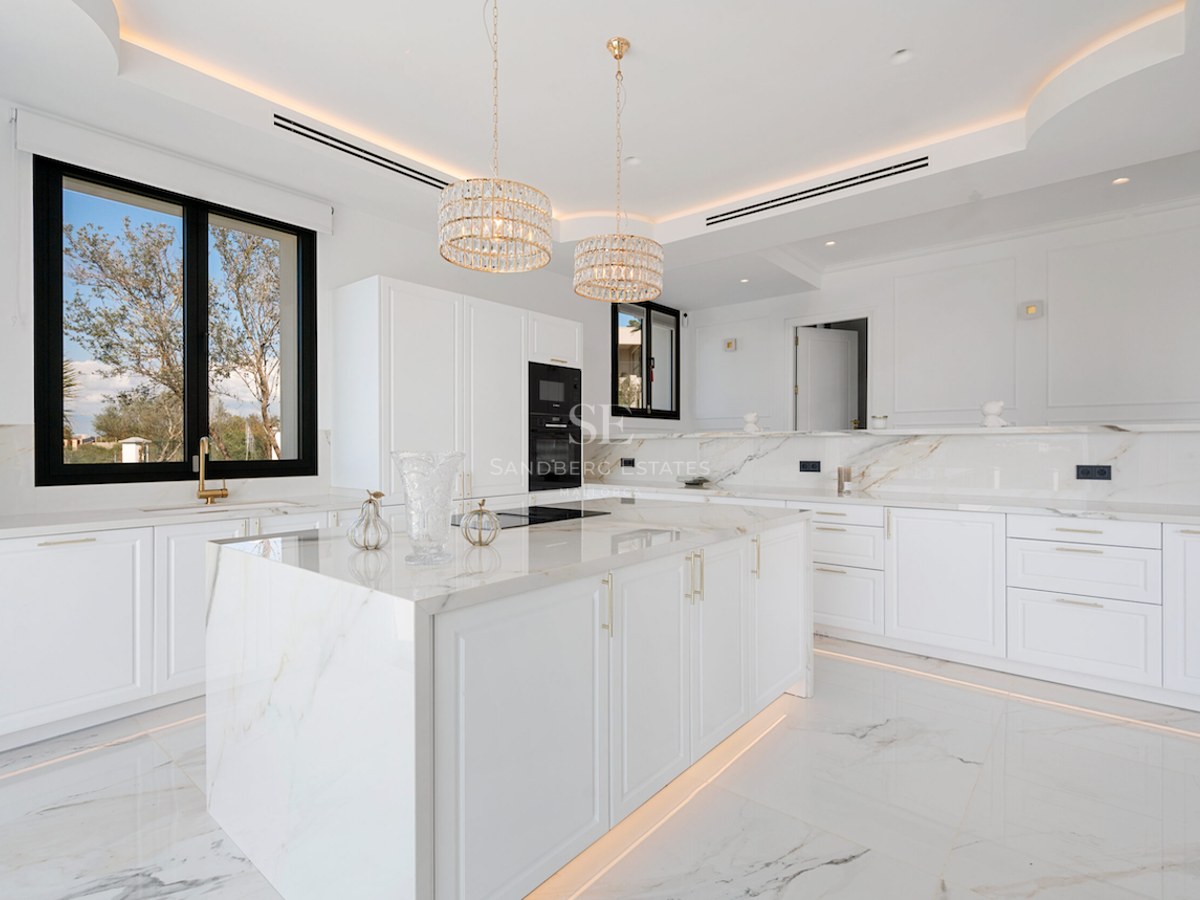 Cocina moderna y luminosa con isla central de mármol, detalles dorados y amplios ventanales.