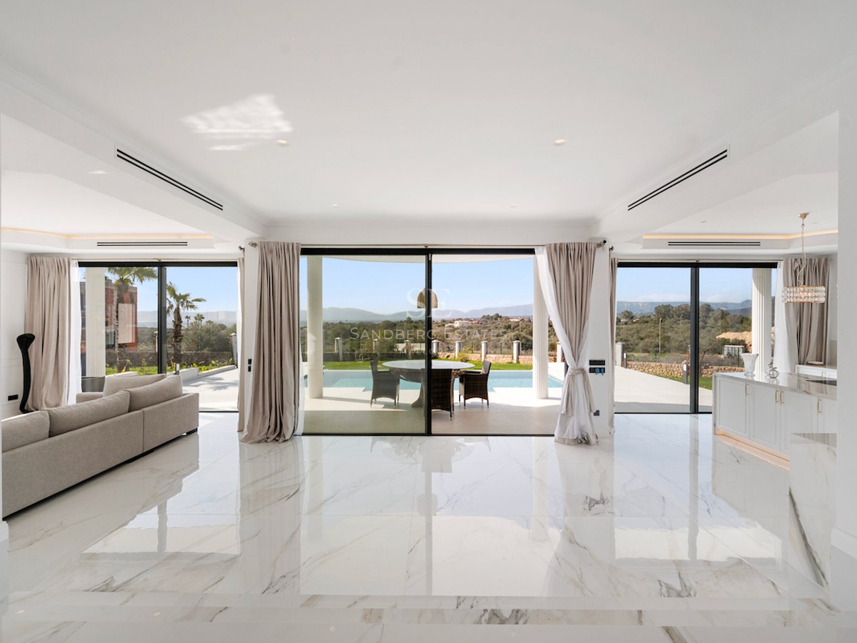 Salón luminoso con suelos de mármol blanco y ventanales de suelo a techo con vistas a la piscina y colinas.