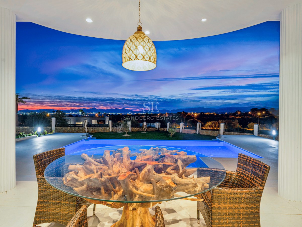 Terraza cubierta con mesa de raíz natural, sillas de mimbre y piscina iluminada con vistas lejanas al atardecer.
