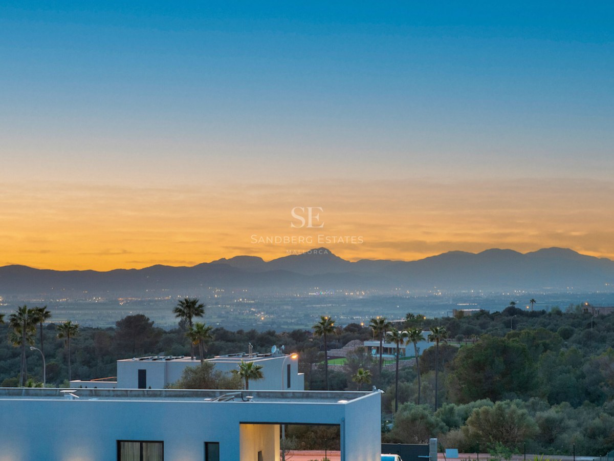 Villa moderna con vistas a un valle y montañas durante un atardecer dorado con luces de la ciudad al fondo.