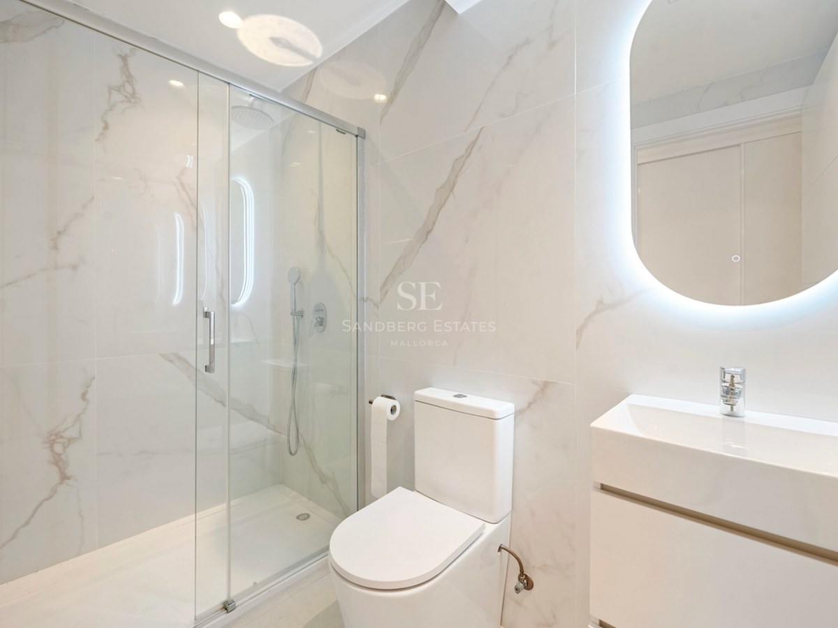 Baño moderno con paredes de efecto mármol, ducha de cristal y espejo ovalado retroiluminado sobre lavabo blanco.