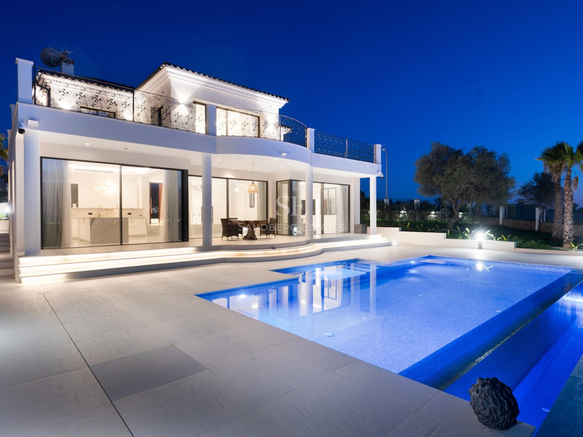 Villa de lujo blanca de dos plantas al atardecer con una gran piscina iluminada, terraza de piedra y balcones de forja.