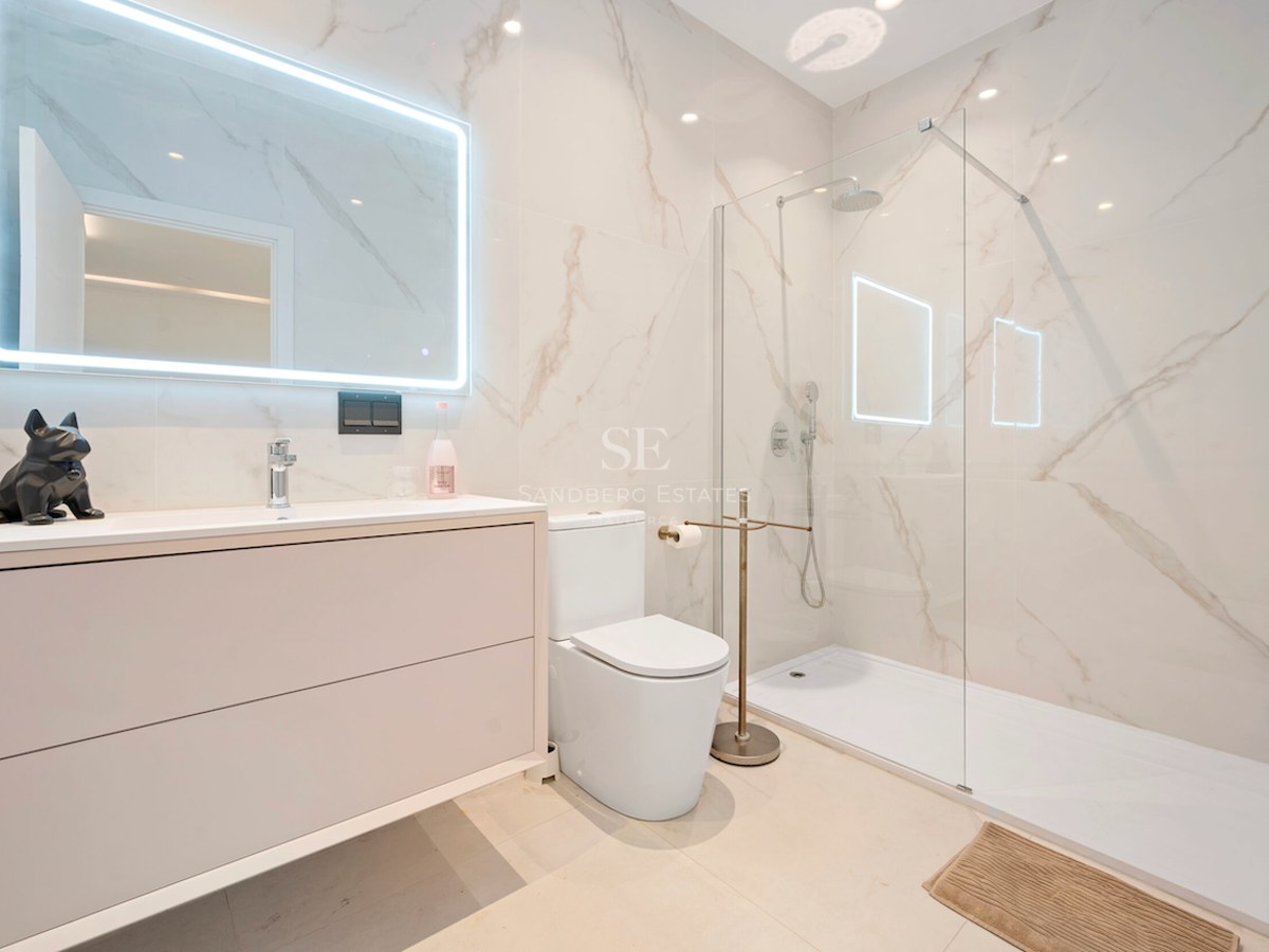 Baño luminoso con ducha de cristal, azulejos de mármol blanco, mueble de lavabo y espejo rectangular retroiluminado.