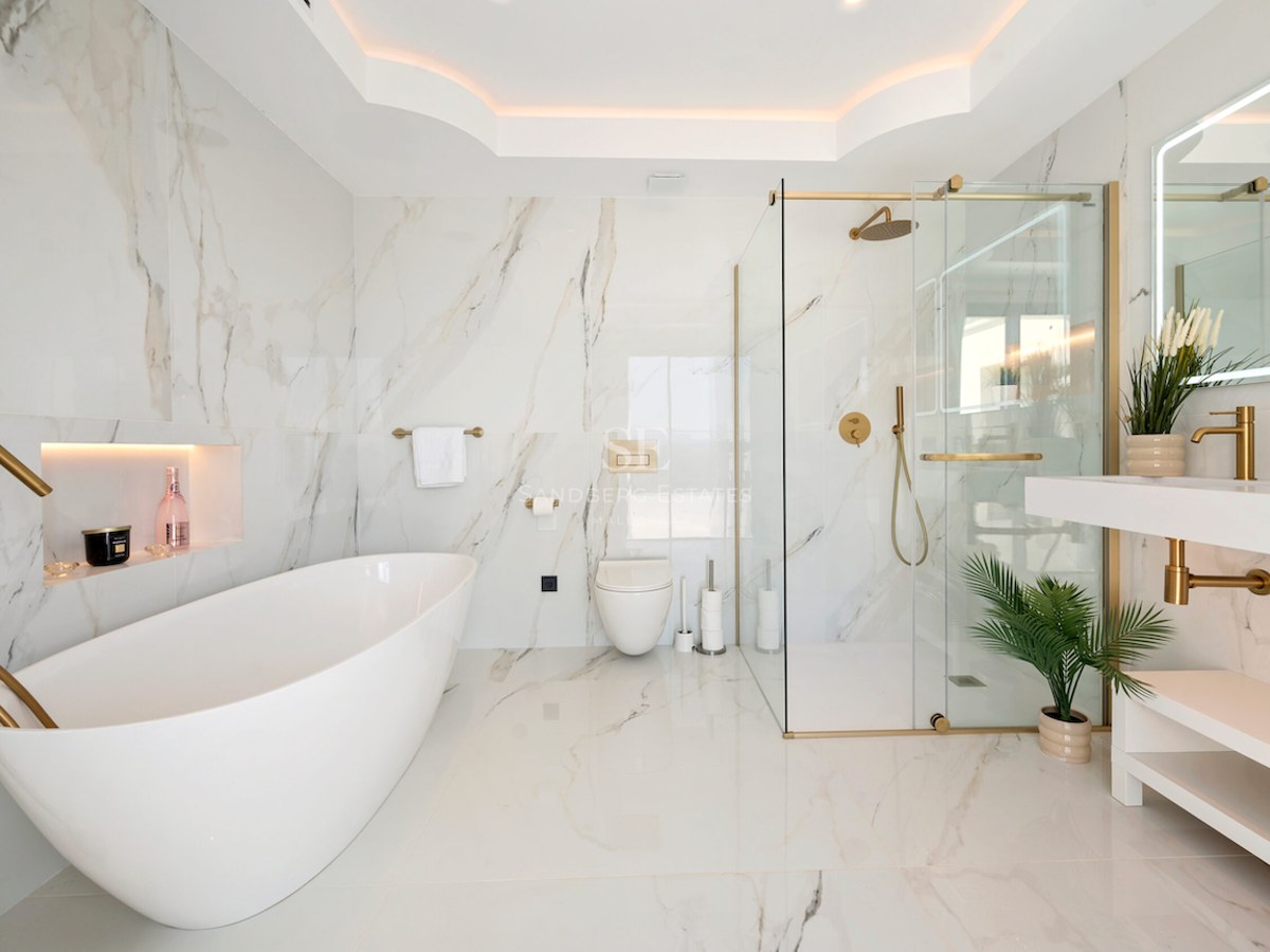 Baño de lujo con bañera exenta blanca, ducha de cristal y grifería dorada sobre paredes de mármol veteado.