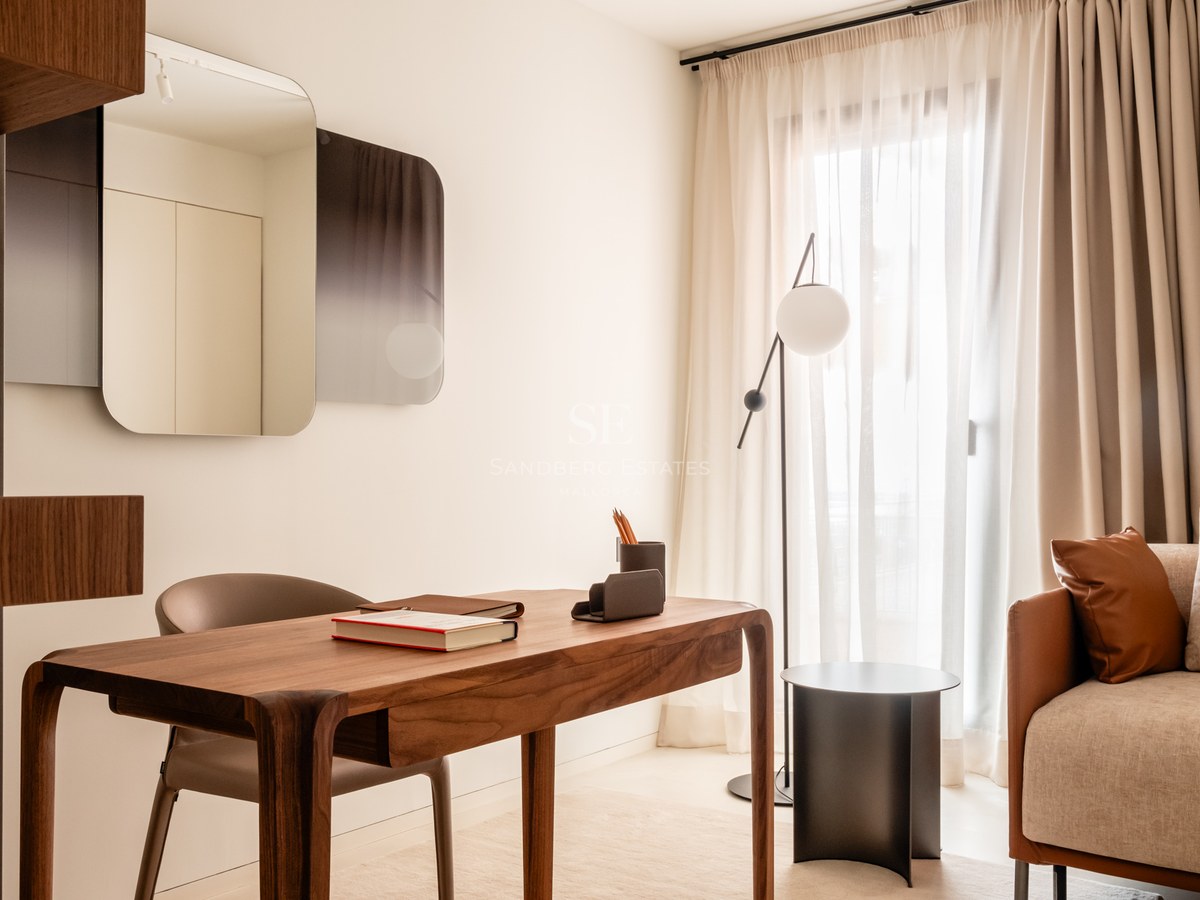 Modern wooden desk and leather chair in a bright room with sheer curtains and a minimalist floor lamp.