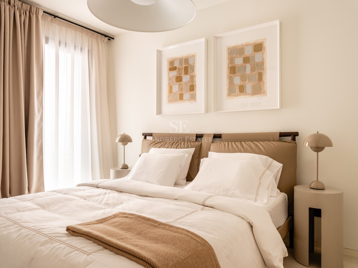 Modern bedroom with a tan leather headboard, white linens, and large windows with sheer curtains.