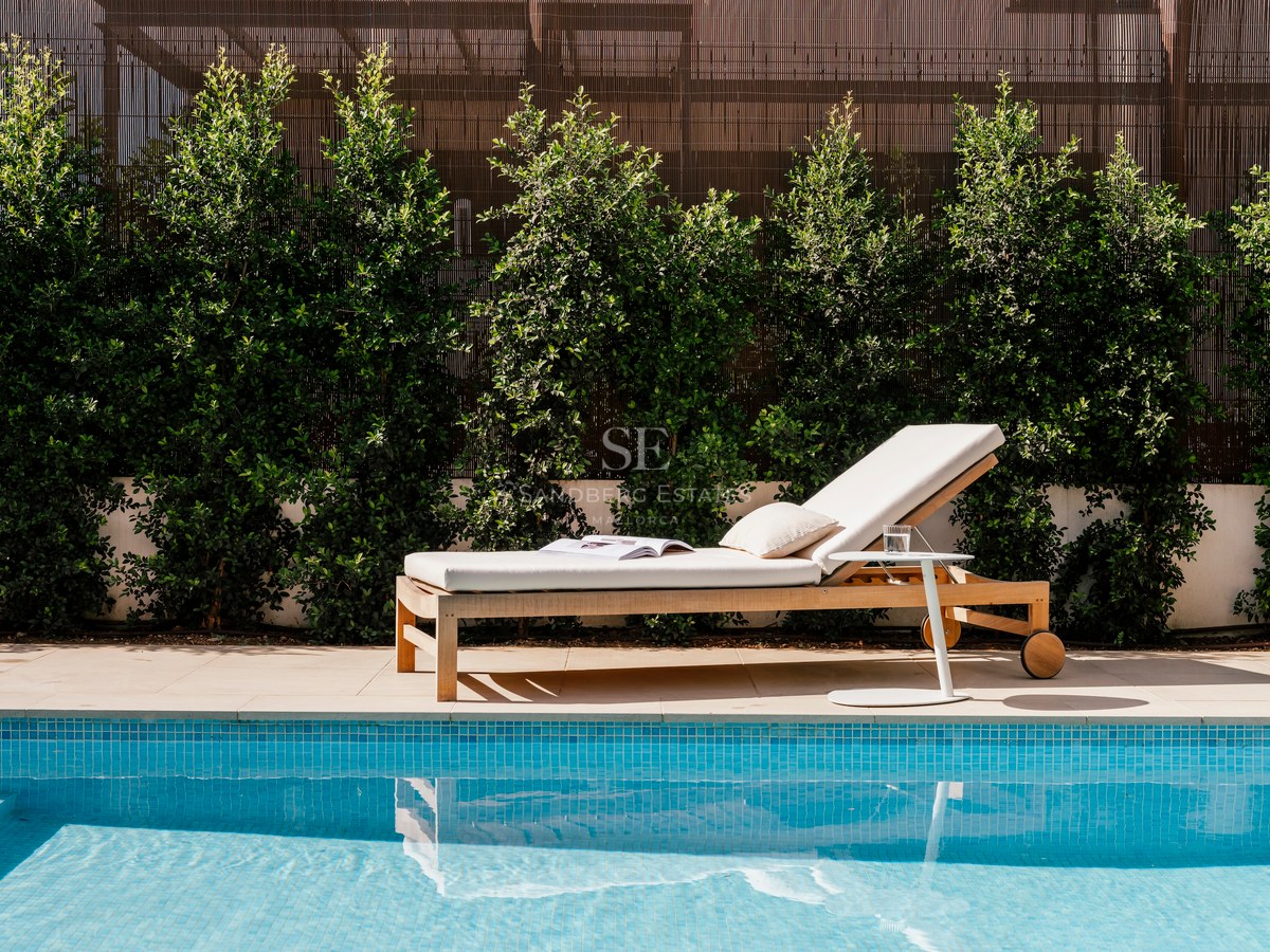 Bain de soleil en bois avec coussins blancs à côté d'une piscine en mosaïque bleue et d'une haie verdoyante.