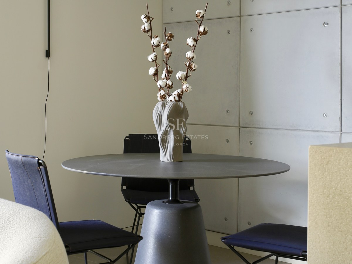 Modern round grey pedestal table with navy chairs against a concrete panel wall and minimalist wall lamp.