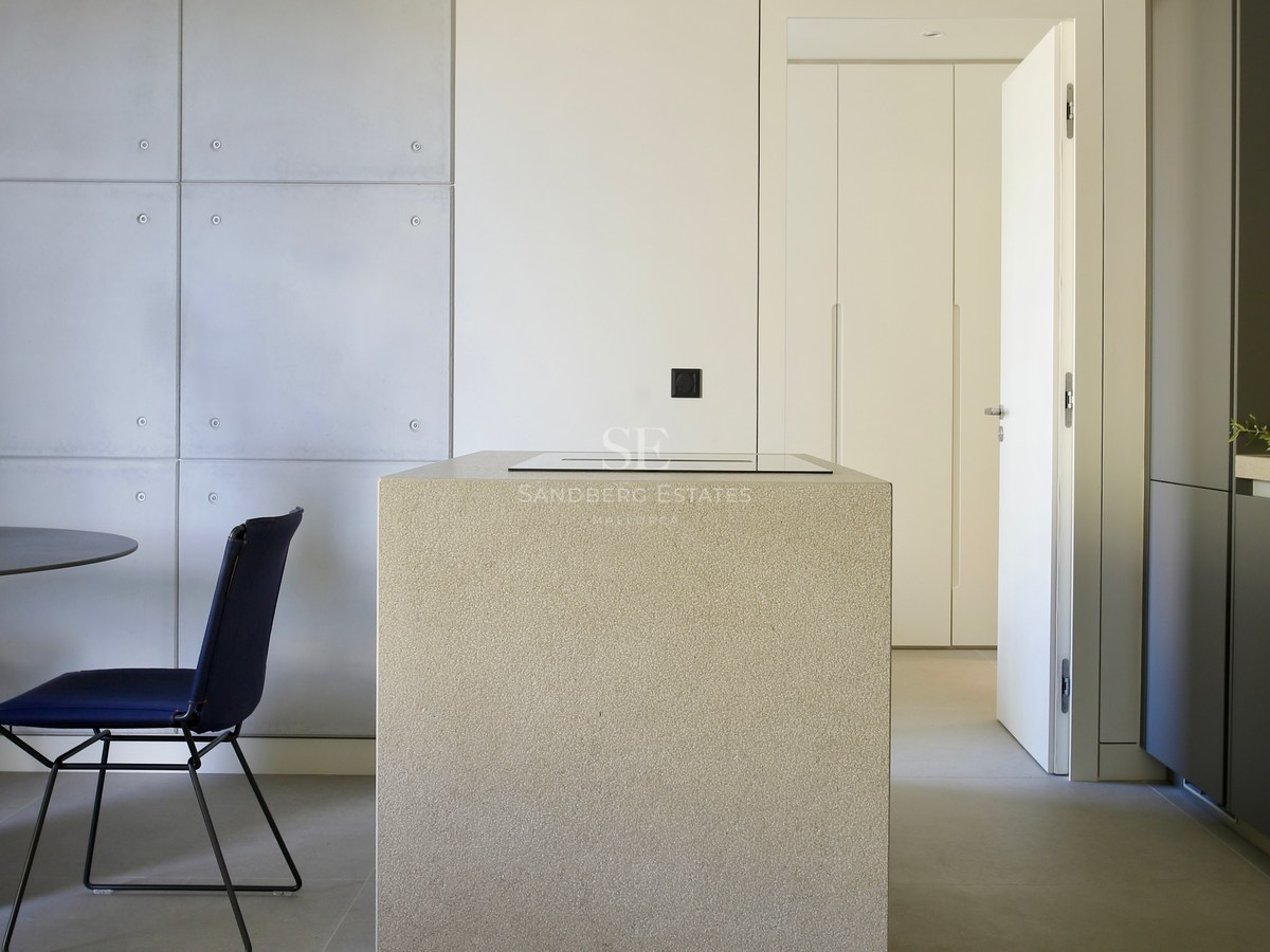 Modern kitchen featuring a textured stone island, concrete wall panels, and a navy blue chair on grey tiled floors.