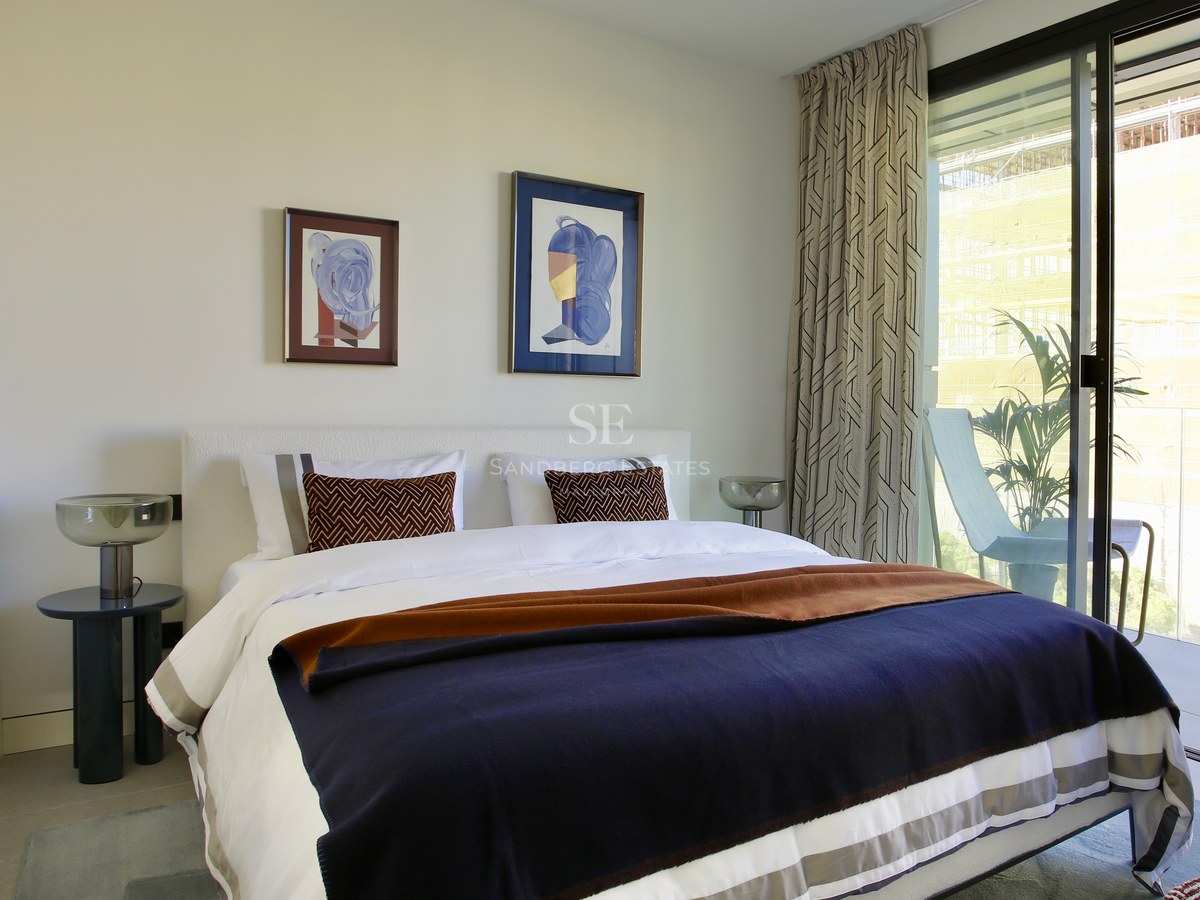 Contemporary bedroom featuring white bedding, navy throw, and floor-to-ceiling glass doors opening to a sunlit balcony.