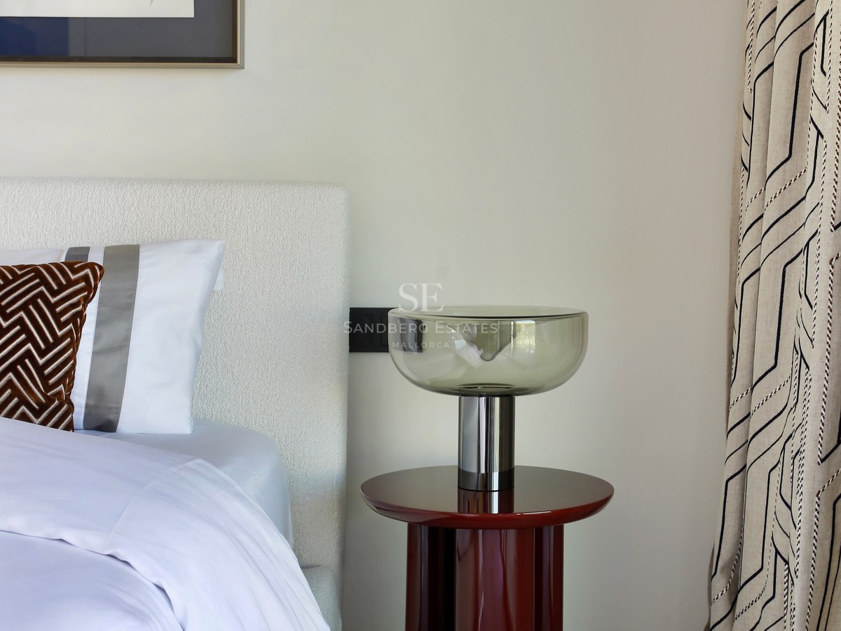 Bedroom detail featuring a white bouclé headboard, red lacquered side table, glass lamp, and abstract art.