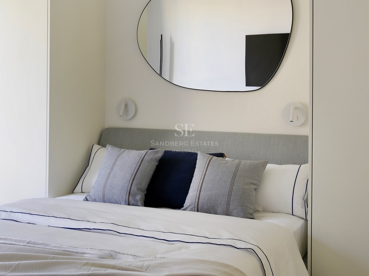 A modern bedroom featuring a neutral-toned bed, an organic-shaped wall mirror, and integrated white wardrobes.