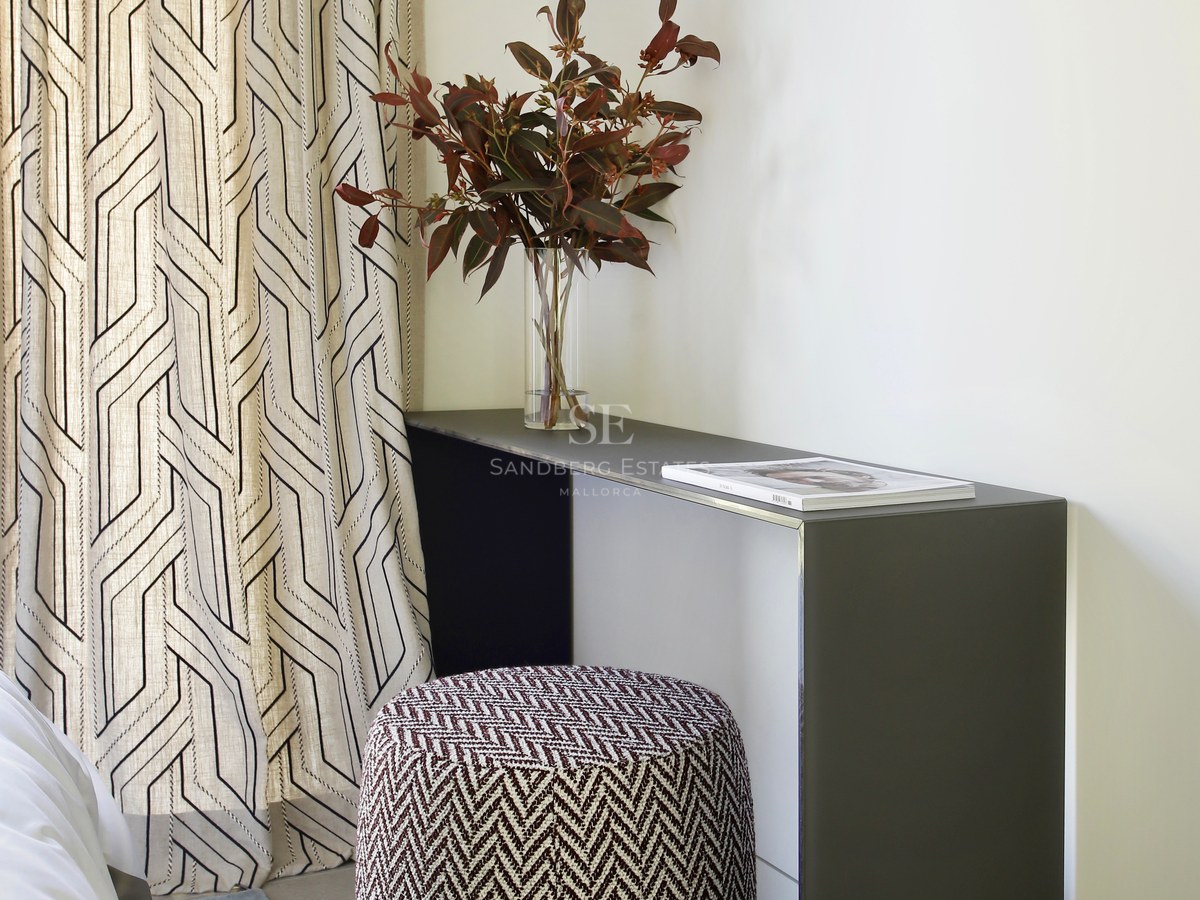 Modern bedroom corner featuring a minimalist gray console, patterned ottoman, and geometric curtains with natural light.