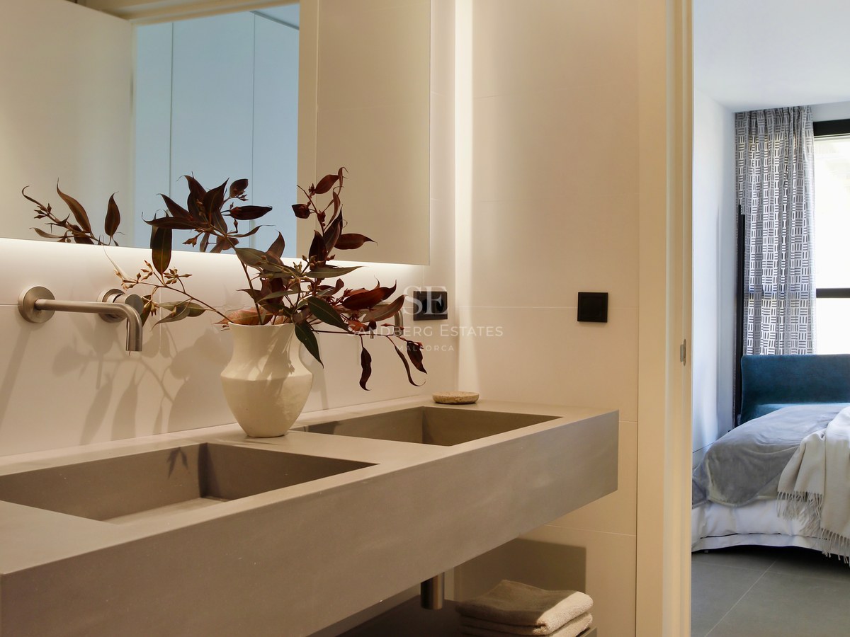 Contemporary bathroom with a grey double sink, large backlit mirror, and view into the adjacent bedroom.