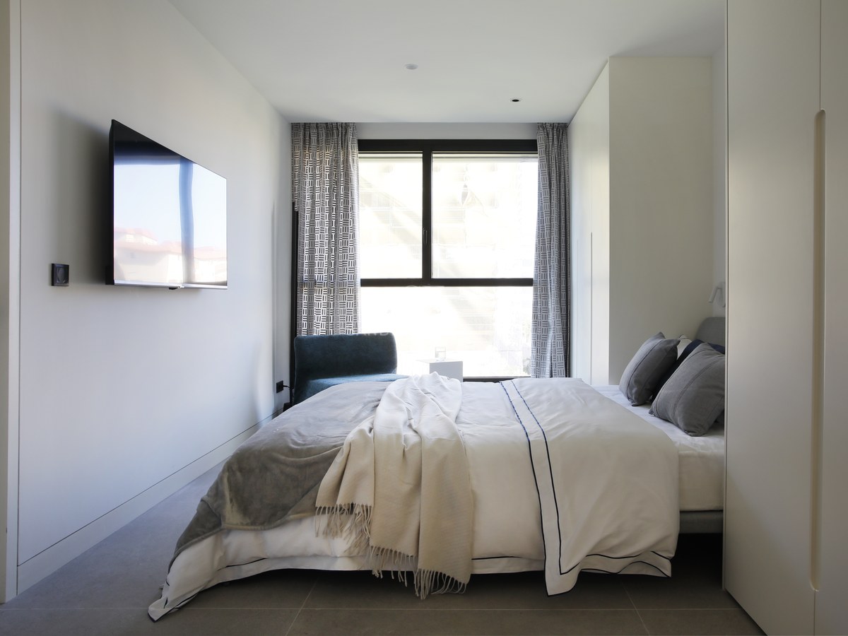 Minimalist bedroom with white walls, a large window, wall-mounted TV, and grey tiled flooring.