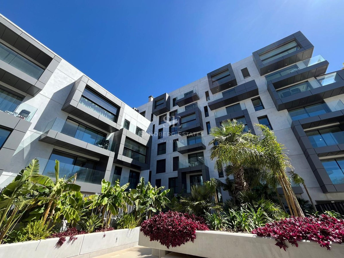 Low-angle view of a modern apartment building with white and grey facade, glass balconies, and tropical landscaping.