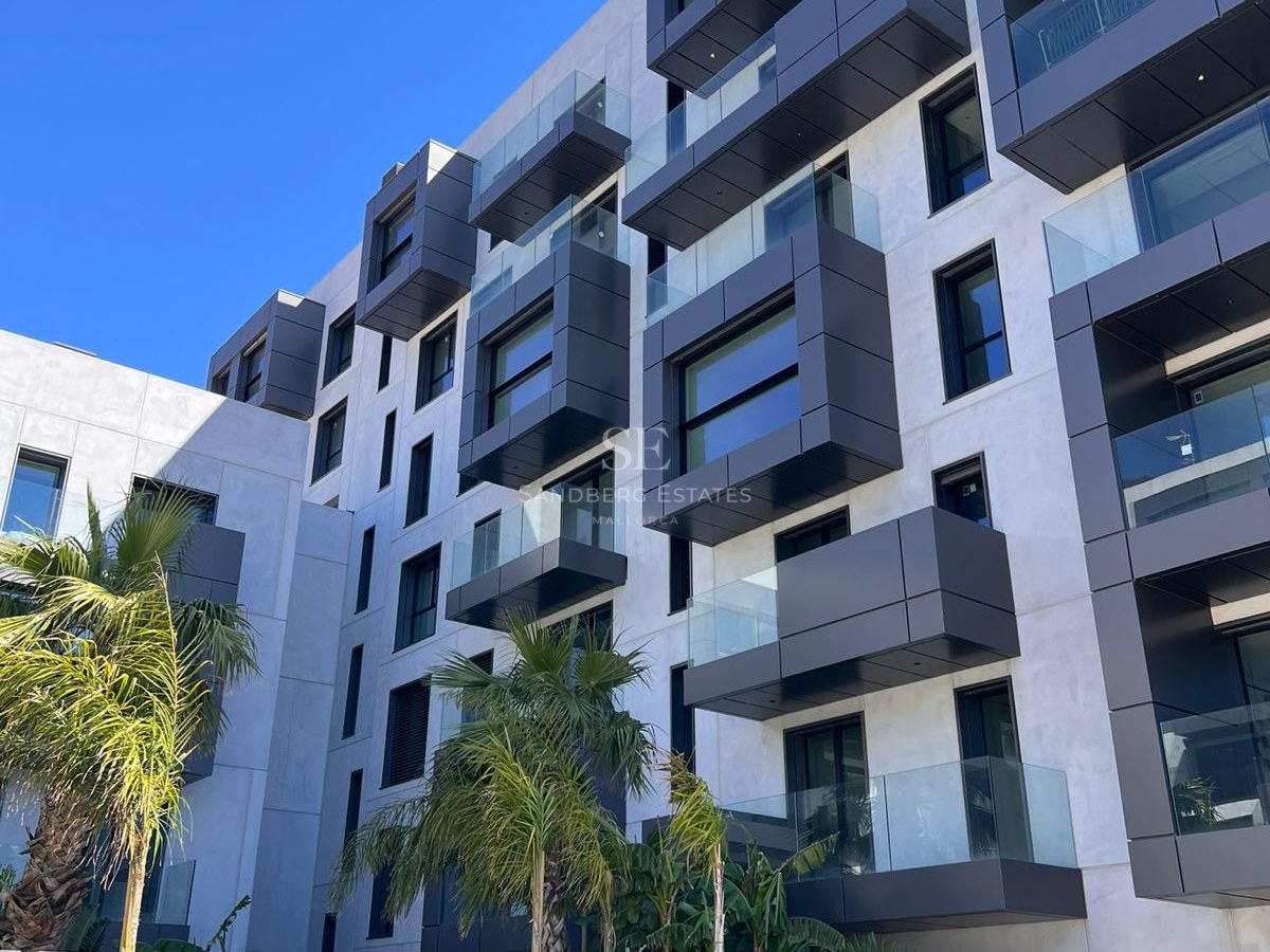 Modern multi-story apartment building with off-white walls, dark gray boxy balconies, glass railings, and palm trees.