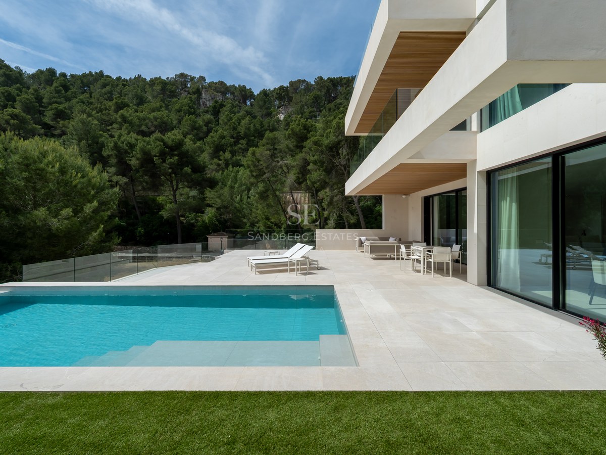 Villa blanca moderna con piscina turquesa, terraza de piedra y barandillas de cristal frente a un bosque de pinos.