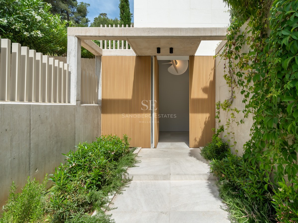 Entrada de villa moderna con listones de madera, muros de hormigón visto y exuberante vegetación mediterránea.