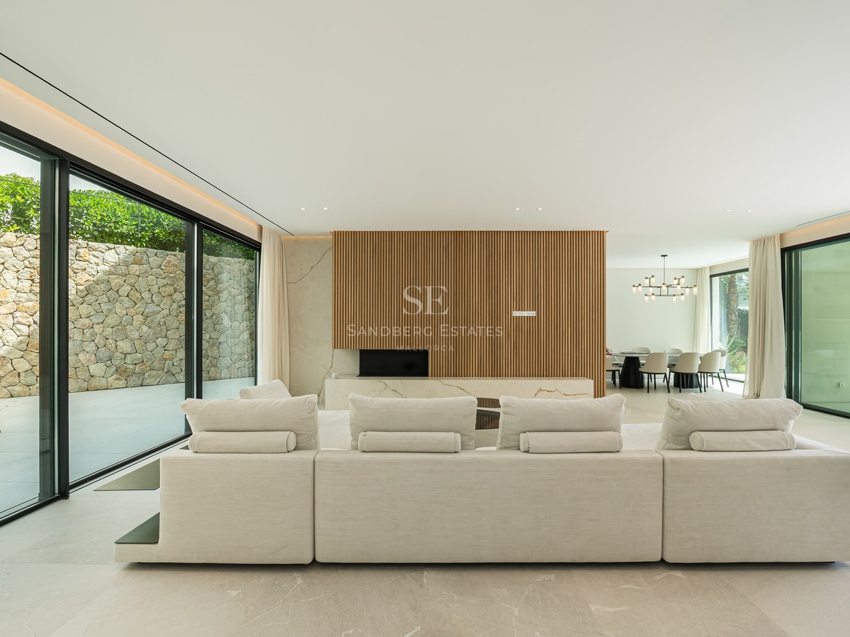 Amplio sofá modular blanco en un salón luminoso con grandes ventanales de cristal y paneles de madera.