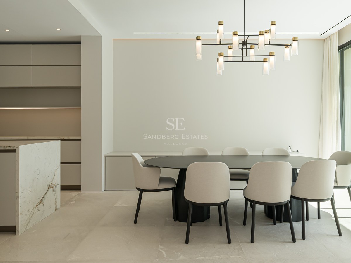 Comedor minimalista con mesa negra, sillas crema, isla de mármol y ventanal de suelo a techo hacia el jardín.