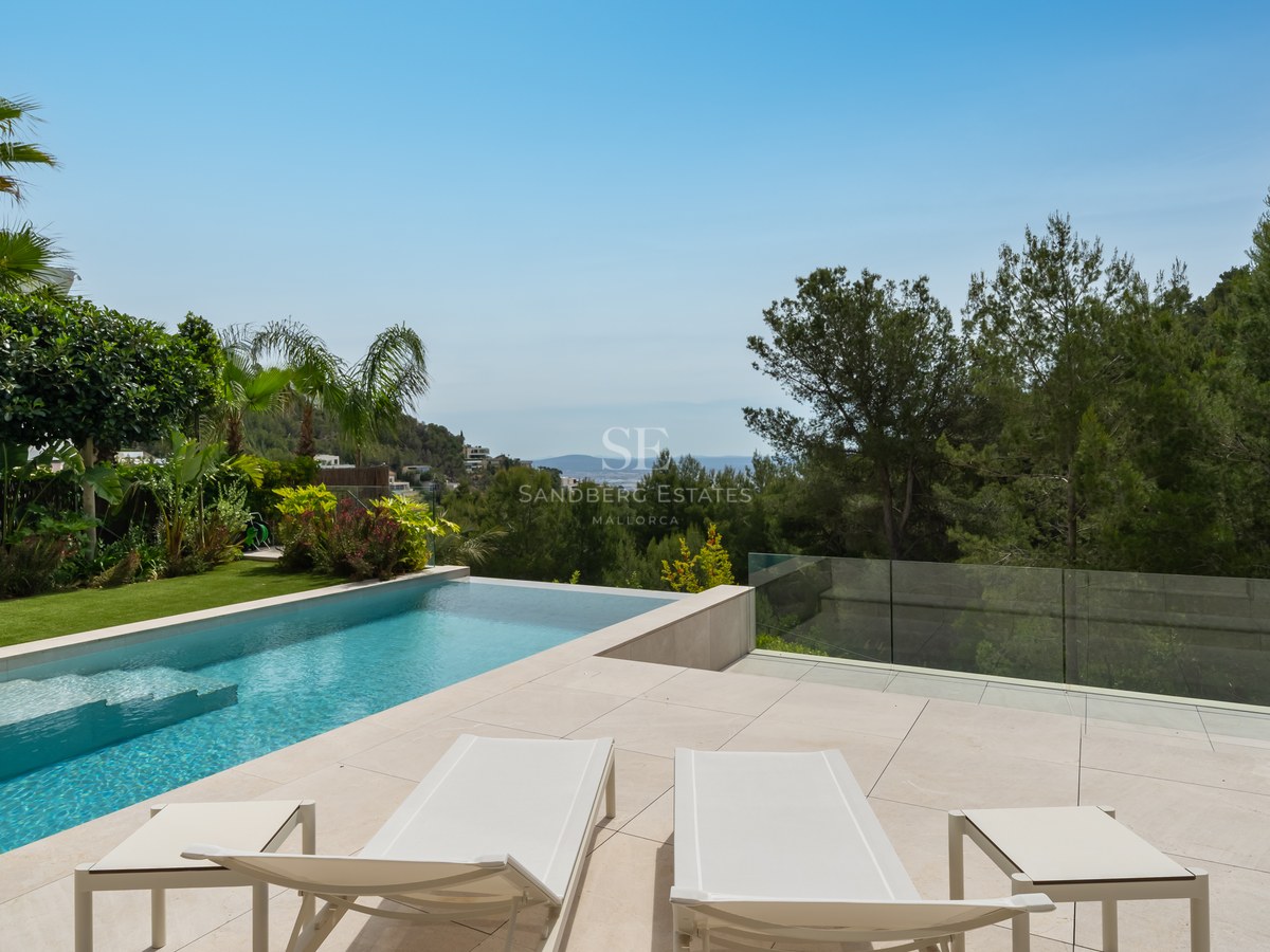 Piscina infinita moderna y terraza de piedra con hamacas blancas frente a un bosque de pinos y el Mediterráneo.