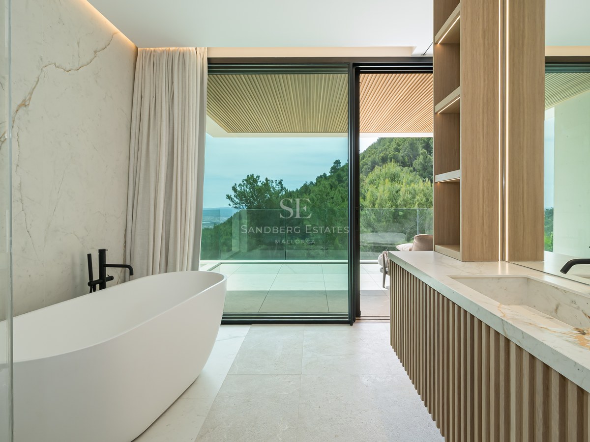 Baño moderno con bañera exenta, paredes de mármol y ventanales con vistas a un bosque de pinos.