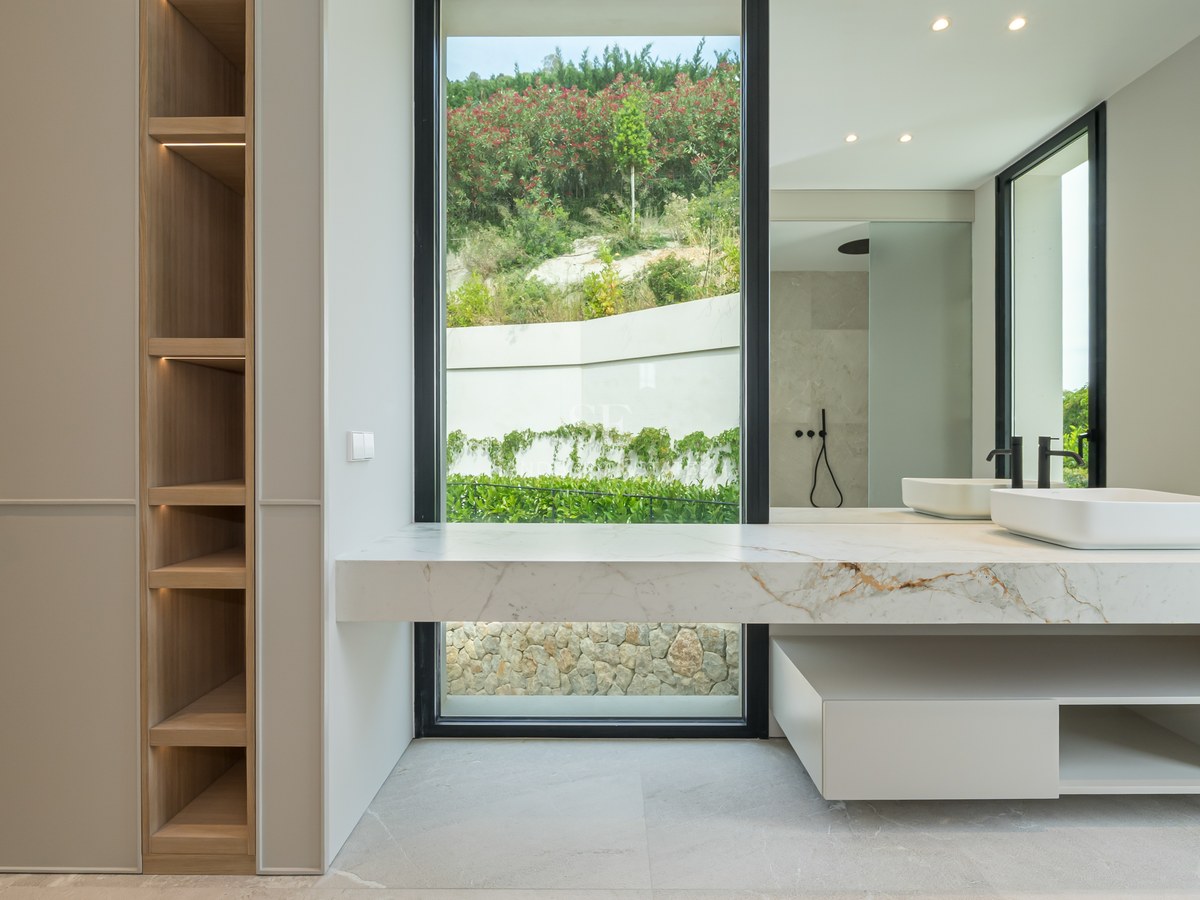 Baño moderno con encimera de mármol, lavabo sobre encimera, grifería negra y ventanal con vistas a la naturaleza.