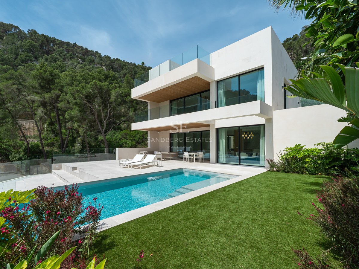 Villa blanca contemporánea con piscina rectangular, césped verde y balcones de cristal frente a una colina boscosa.