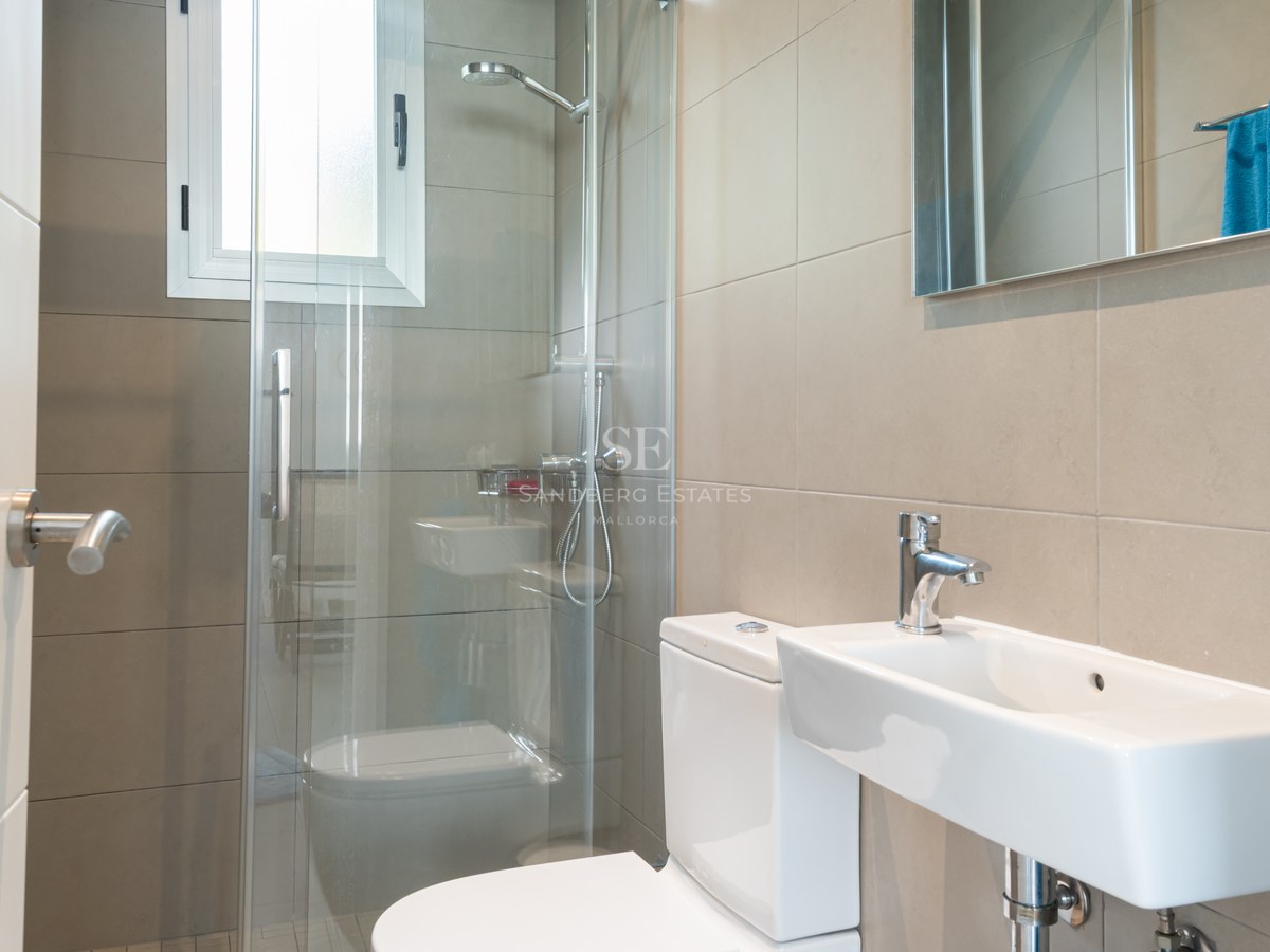 Baño contemporáneo con azulejos beige, ducha con mampara de cristal, lavabo blanco y luz natural.