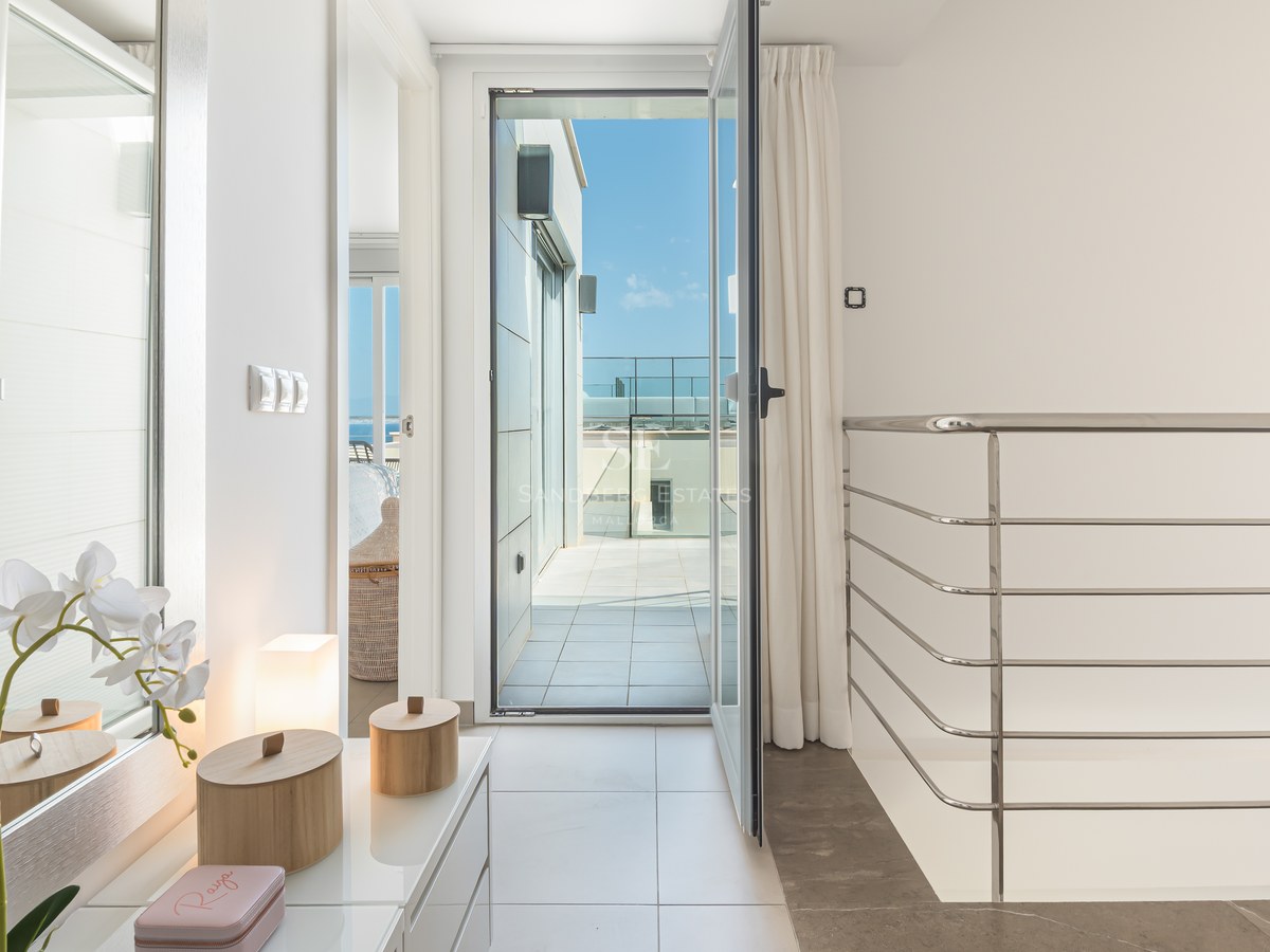 Ljus hall med vitt klinkergolv som leder till en öppen glasdörr och en solig terrass med metallräcke.