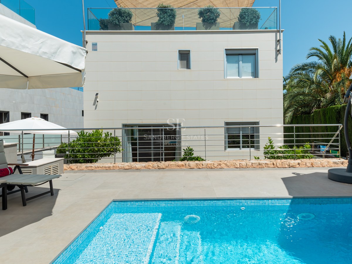 Privat utomhuspool med turkos mosaik, solstolar och vita parasoll framför en modern villafasad.