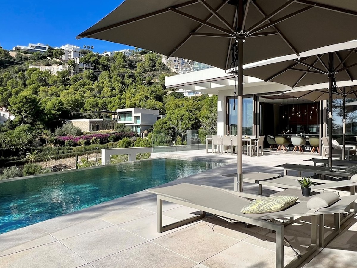 Vista exterior de villa moderna con piscina infinita, amplios ventanales y jardines exuberantes. Diseño minimalista y elegante.