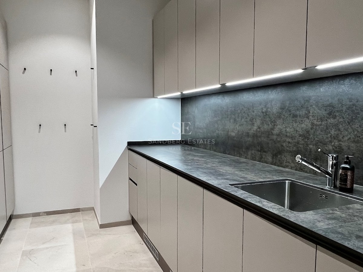 Lavandería moderna con encimera de piedra gris, muebles beige sin tiradores, iluminación LED y suelo de piedra.