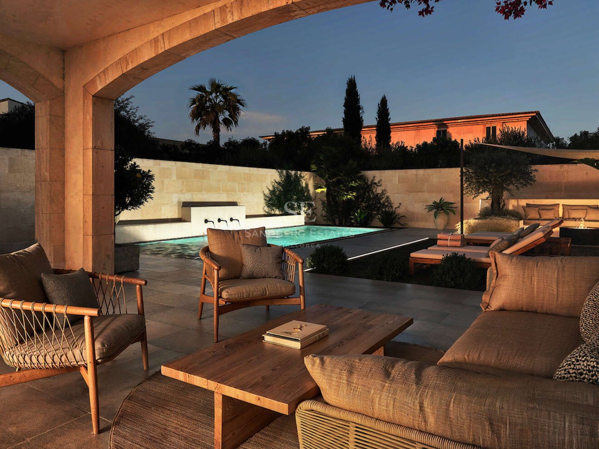 Salon extérieur avec mobilier en bois donnant sur une piscine illuminée et un jardin au coucher du soleil.