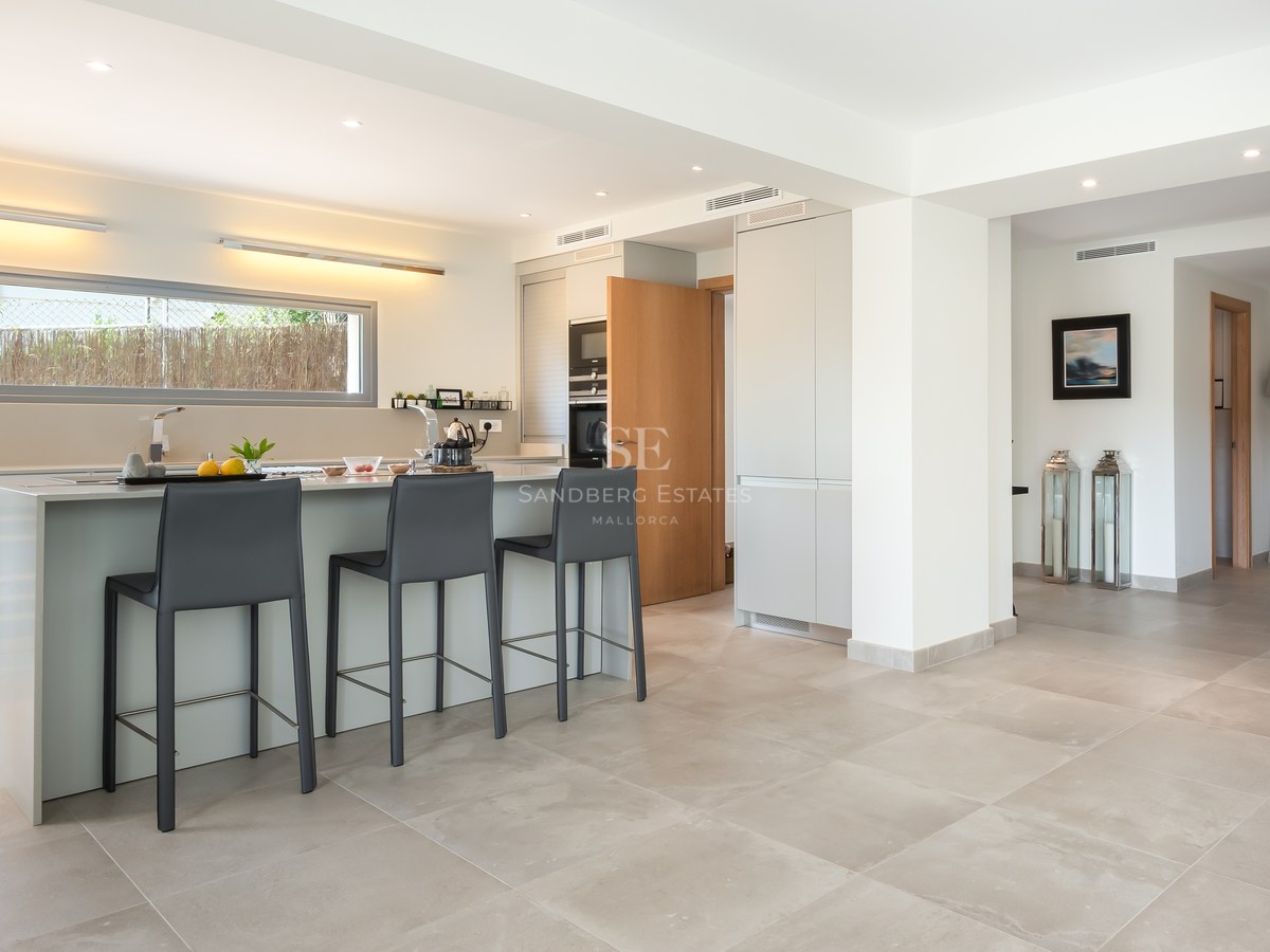 A contemporary kitchen featuring a large light grey island with three charcoal bar stools, a wine cooler, and tile flooring.