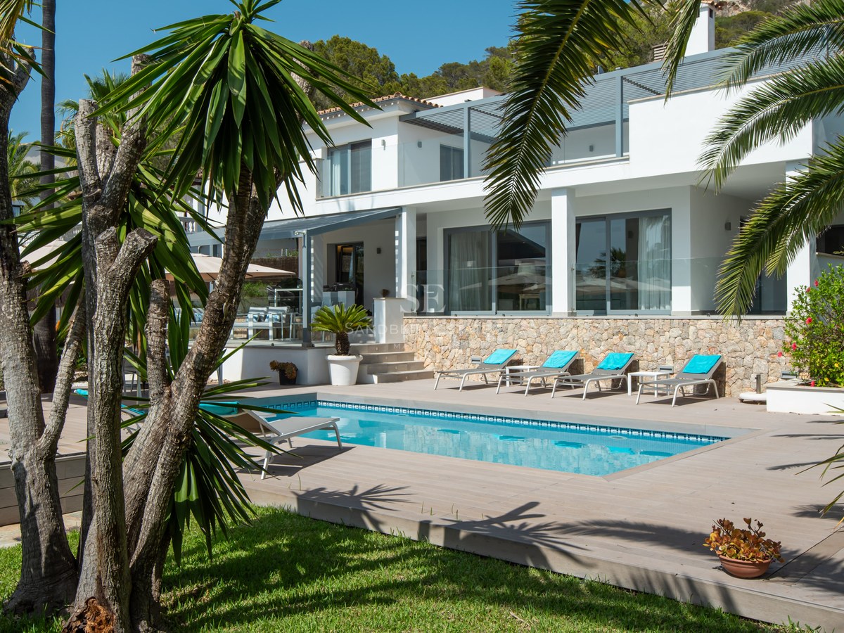 Modern white villa with a rectangular swimming pool, sun loungers, and lush palm trees under a clear blue sky.